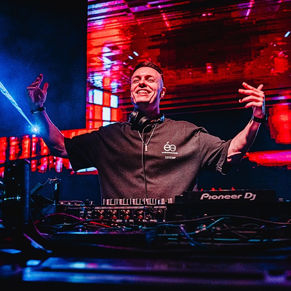 Tiscore smiling and performing live behind the DJ booth with vibrant red visuals in the background.